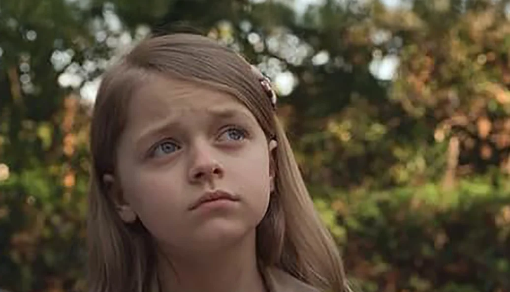 McKenna Grace: Close-up de uma menina loira com olhos claros olhando para cima com expressão melancólica ou contemplativa.