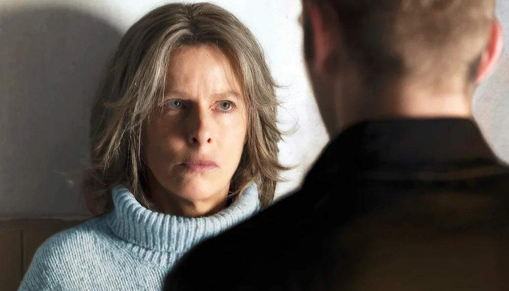 Karin Viard: Uma mulher de cabelos grisalhos e gola alta azul olha intensamente para um homem que está de costas em primeiro plano.