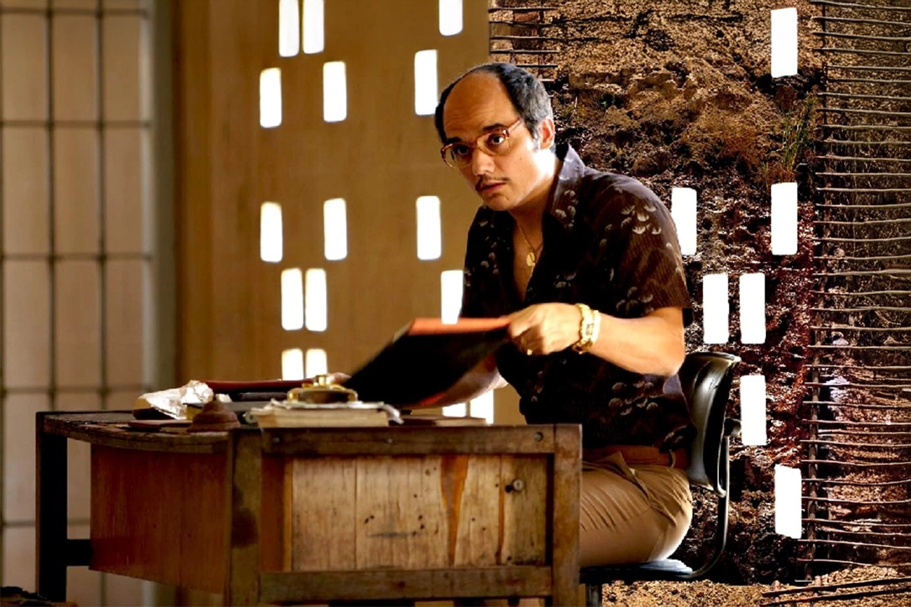 Caio Blat: Homem calvo de óculos e bigode sentado à uma mesa de escritório em um ambiente rústico, segurando uma pasta ou livro.