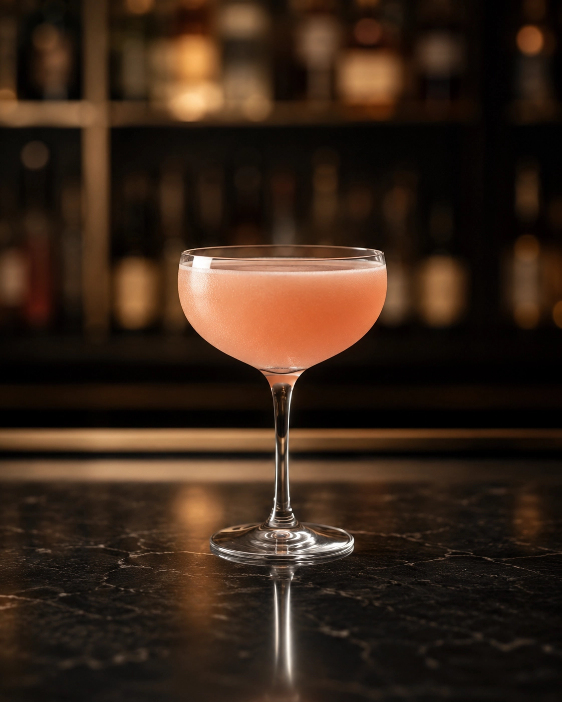 A pink cocktail in a coupe glass on a dark marble bar counter with a blurred bottle display in the background.
