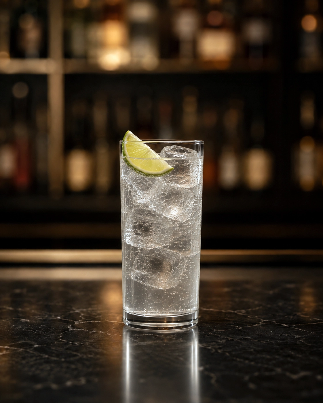 A tall glass containing a clear carbonated drink with ice cubes and a lime wedge garnish, set on a dark marble bar counter with a blurred background of liquor bottles.