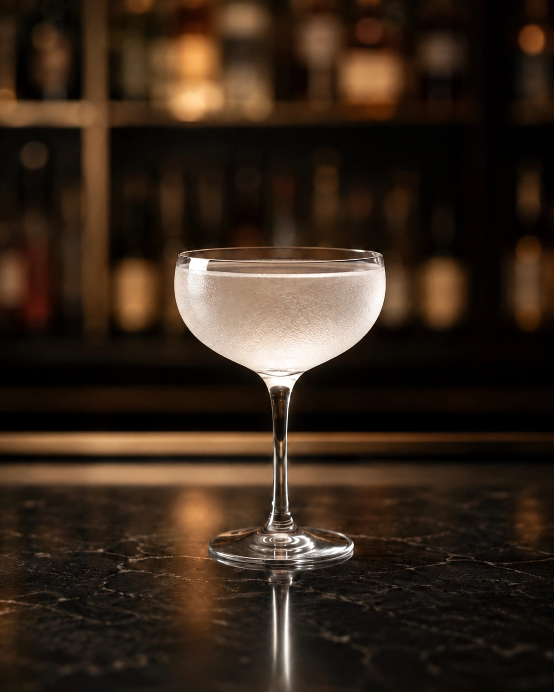 unknown: A close-up shot of a translucent cocktail in a coupe glass on a dark marble bar counter with a blurred background of spirit bottles.
