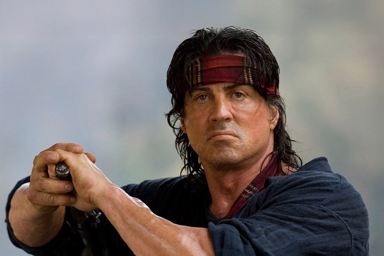 Sylvester Stallone: Fotografia de Sylvester Stallone caracterizado como o personagem John Rambo, usando uma bandana vermelha e segurando uma arma.