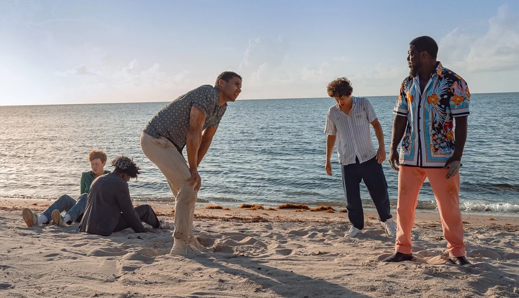 Kevin Hart: Uma cena do filme 'On Our Way' (ou similar) mostrando um grupo de homens em uma praia, com o mar ao fundo.
