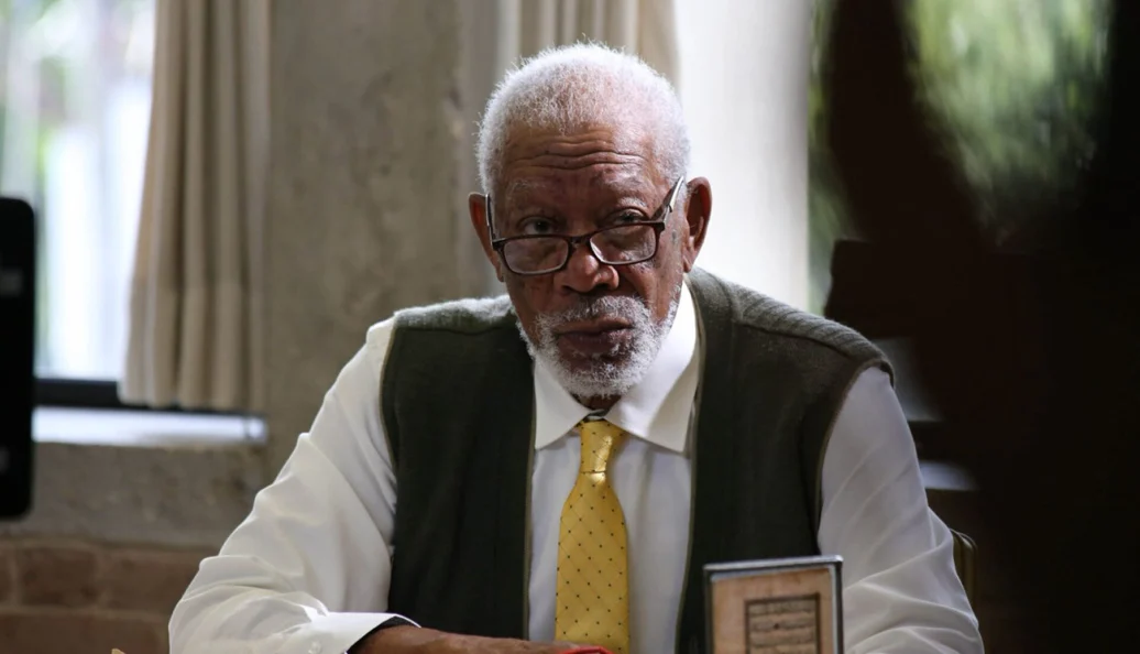 Morgan Freeman: Retrato de Morgan Freeman usando óculos, camisa social branca, gravata amarela e colete de lã, sentado em uma mesa.