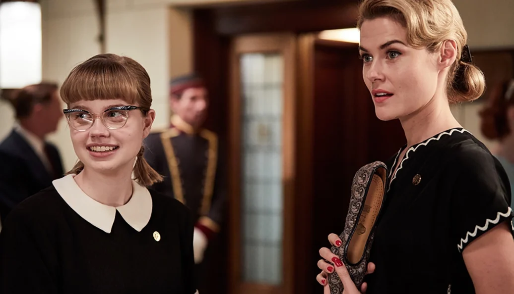 Angourie Rice, Rachael Taylor: Uma cena de filme mostrando duas mulheres em trajes de meados do século XX, uma jovem com óculos sorrindo e outra mulher loira segurando um sapato.