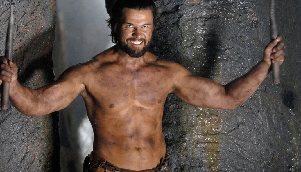 Manu Bennett: Homem musculoso e barbudo, sem camisa, segurando estacas de metal em um ambiente rochoso, possivelmente uma cena de filme ou série.