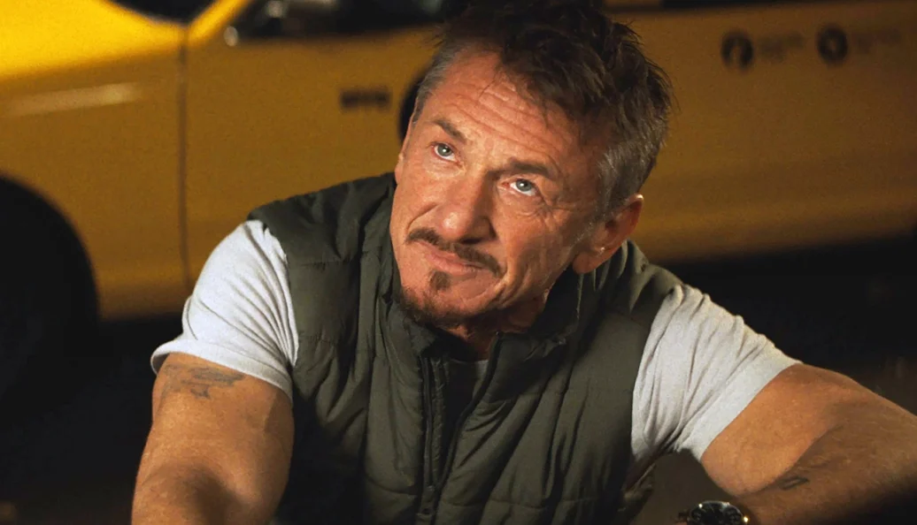 Sean Penn: Close-up de Sean Penn em uma cena de filme, vestindo um colete verde sobre uma camiseta branca, com um carro amarelo ao fundo.