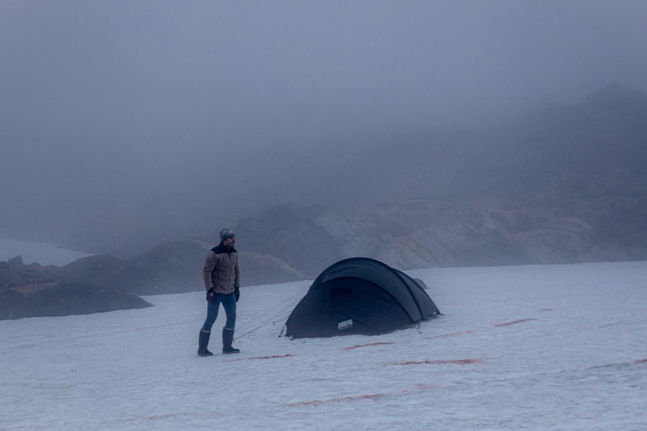Um homem de pé ao lado de uma barraca de acampamento em um terreno coberto de neve sob um céu nublado e enevoado.