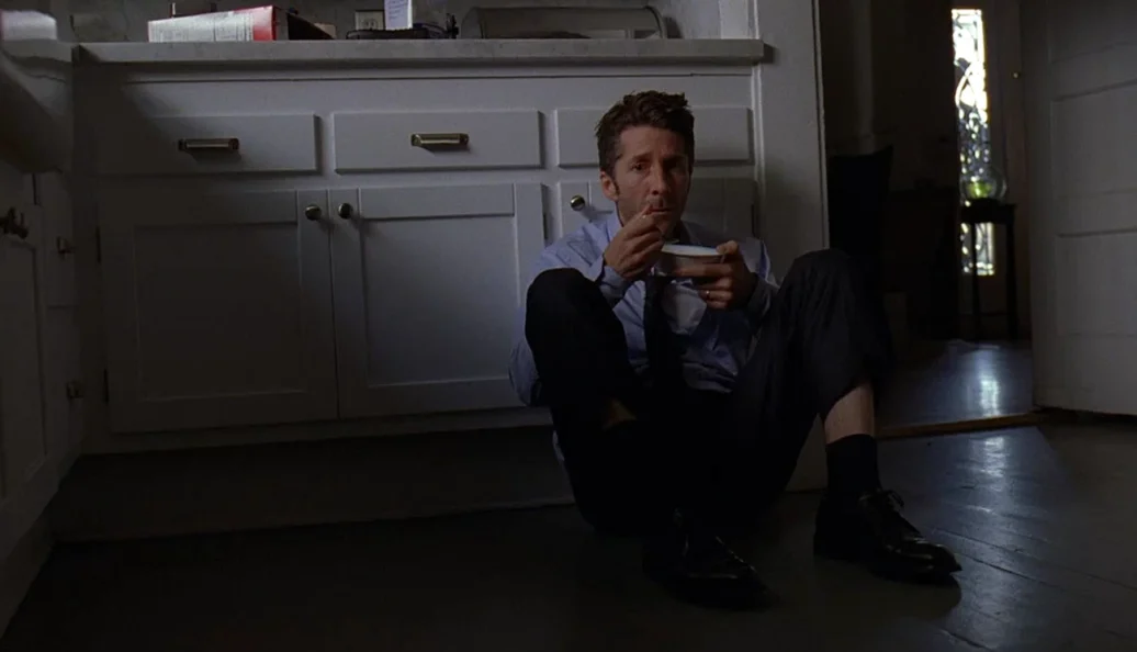 Leland Orser: Homem sentado no chão de uma cozinha, encostado nos armários, comendo em uma tigela. Cena do filme 'O Sexto Sentido'.