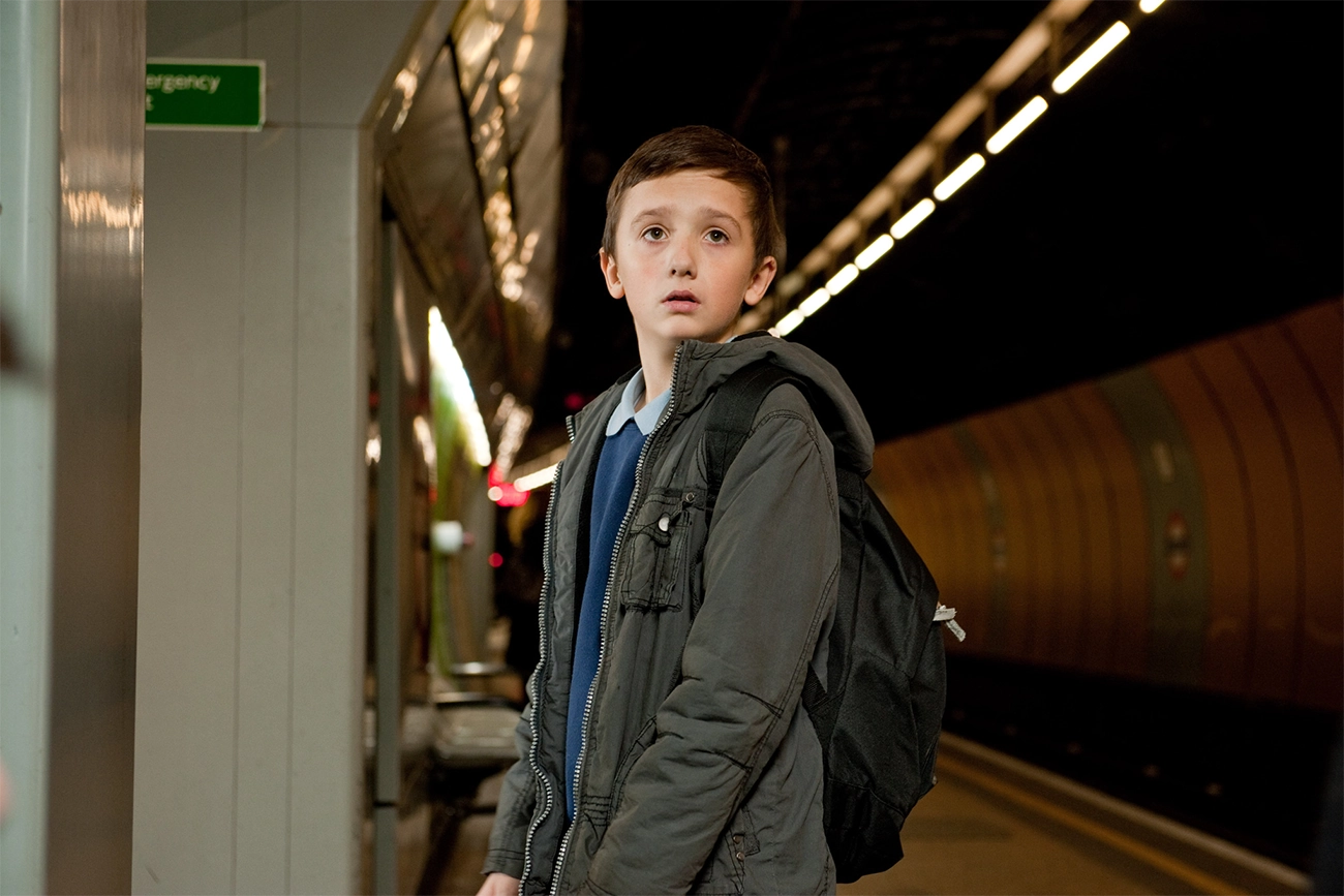 Samuel Joslin: Um menino jovem com uma mochila em pé em uma plataforma de metrô, com uma expressão de incerteza ou preocupação.