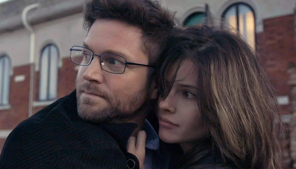 Jason Priestley, Agam Darshi: Um homem de óculos e uma mulher abraçados olhando para a mesma direção, em frente a um edifício de tijolos.