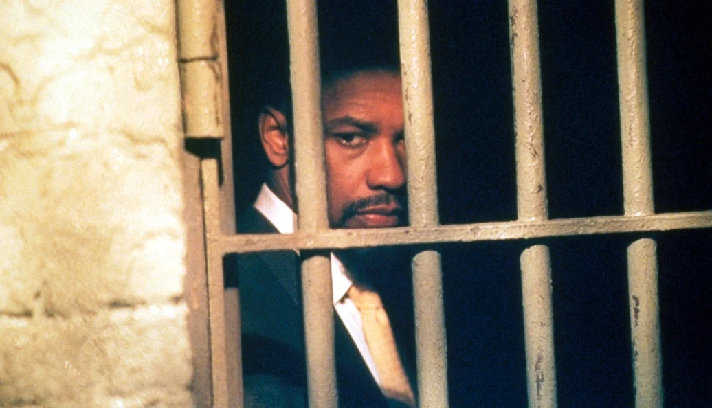 Denzel Washington: Homem negro de terno atrás de grades de uma cela, em cena do filme The Hurricane.