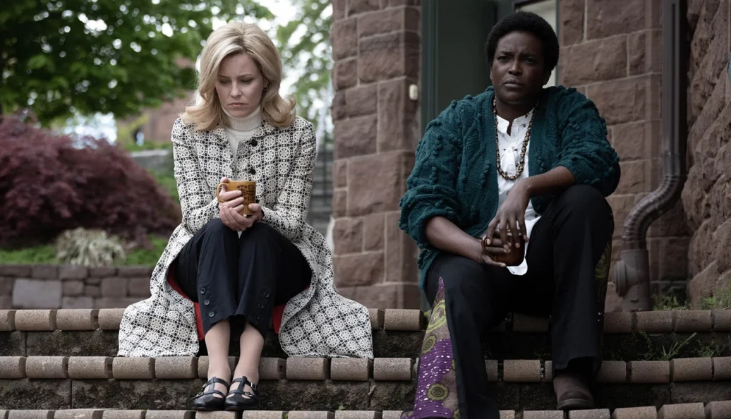 Elizabeth Banks, Wunmi Mosaku: Duas mulheres sentadas em degraus de pedra do lado de fora de uma casa, ambas segurando canecas e parecendo pensativas.