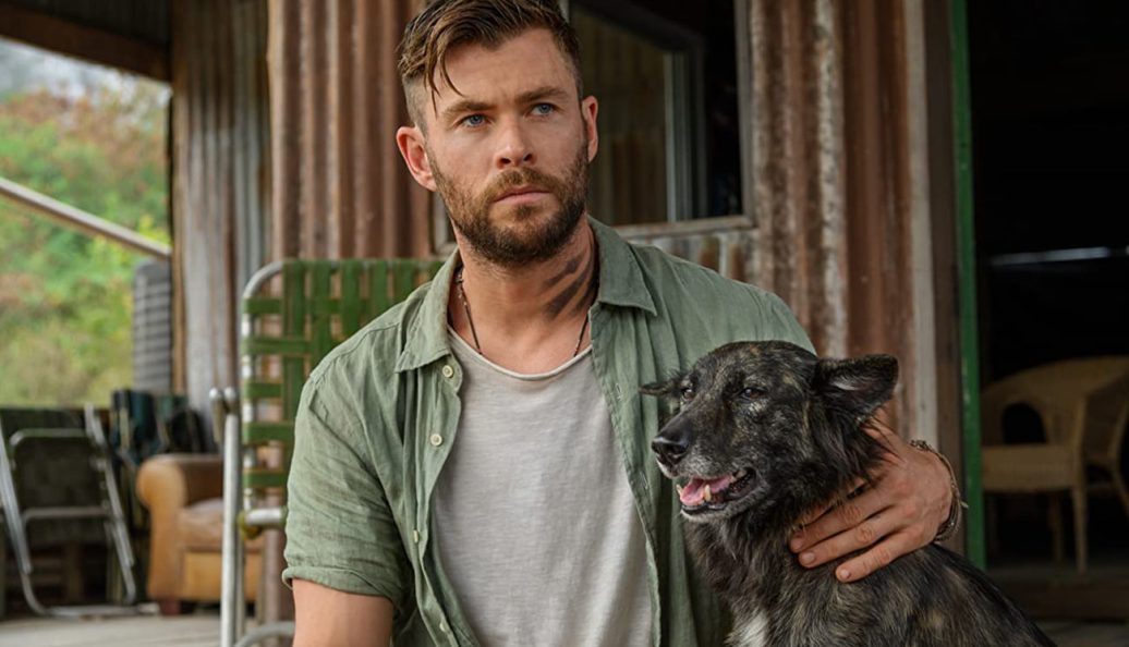 Chris Hemsworth: O ator Chris Hemsworth em uma cena de filme, segurando um cachorro e olhando para o lado.