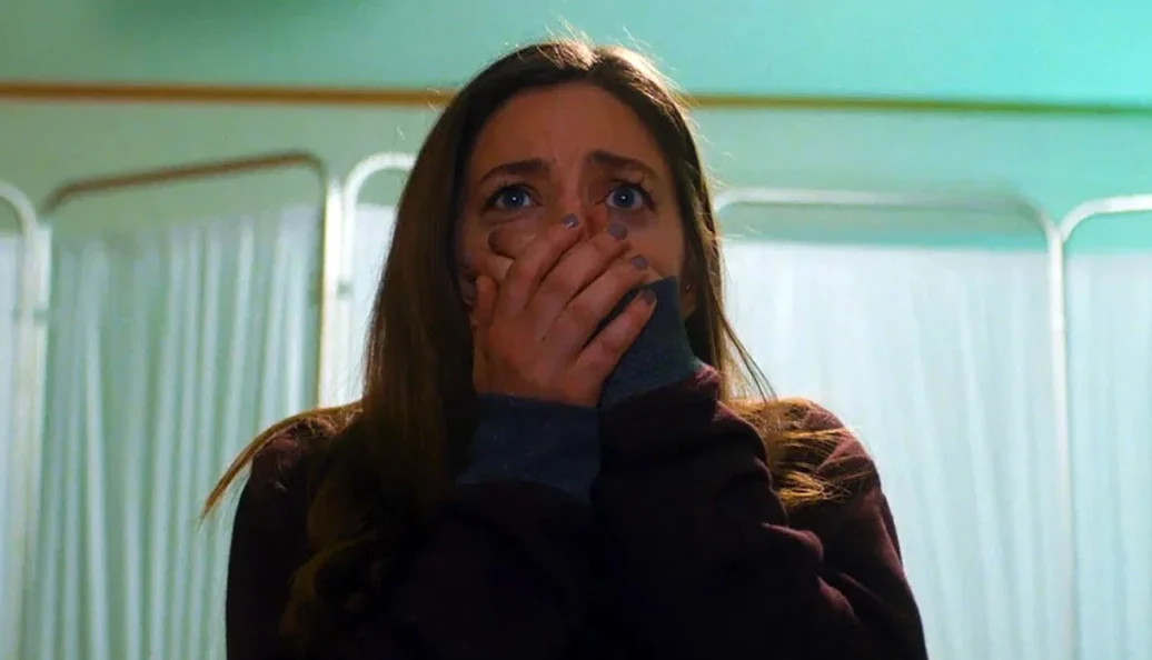 Jessica Rothe: Uma mulher jovem com expressão de choque e pavor, cobrindo a boca com as mãos em um ambiente que parece ser um hospital.