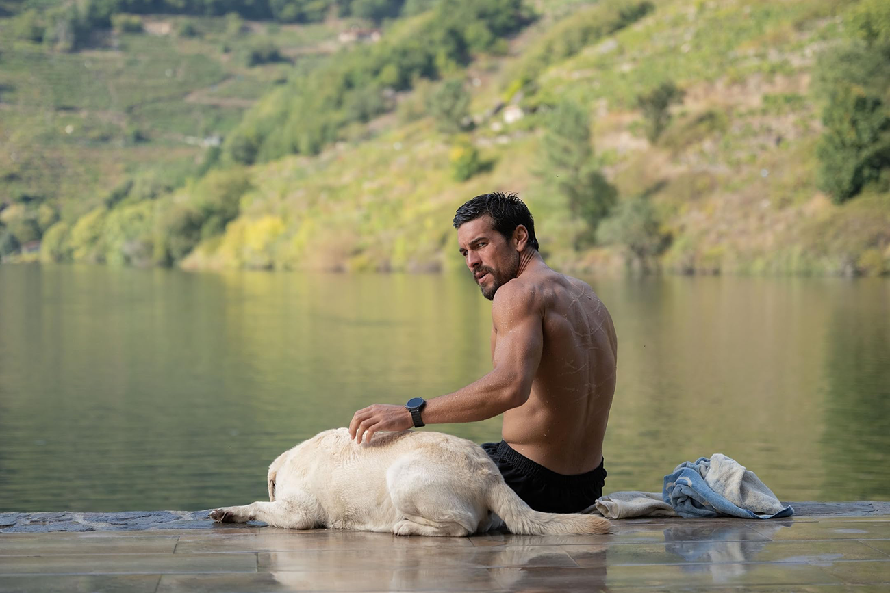 António Félix da Costa: Um homem musculoso sem camisa sentado à beira de um lago ou rio, acariciando um cachorro labrador deitado ao seu lado. Ao fundo, uma encosta com vegetação.