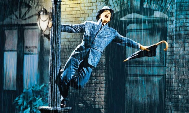 Gene Kelly: Cena clássica do filme musical Cantando na Chuva (Singin' in the Rain) com o protagonista segurando um guarda-chuva e apoiado em um poste sob a chuva.