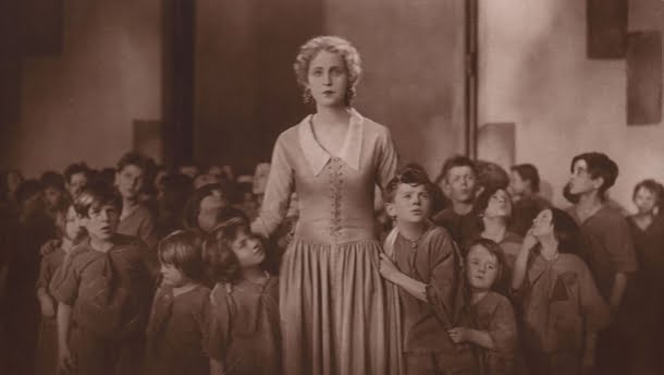 Brigitte Helm: Uma mulher loira em um vestido de época cercada por um grupo de crianças em um cenário que parece ser de um filme clássico.