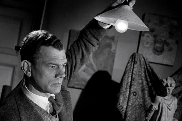 Joseph Cotten: Fotografia em preto e branco de um homem segurando uma luminária acesa para cima, observando algo fora de campo em um ambiente artístico.