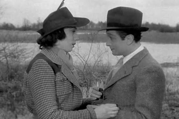 Mila Parély, Roland Toutain: Uma cena em preto e branco de um filme clássico mostrando um homem e uma mulher conversando ao ar livre, ambos usando chapéus e casacos de lã.
