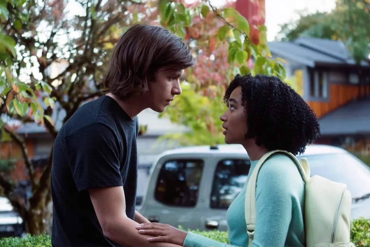 Nick Robinson, Amandla Stenberg: Uma fotografia de cena do filme 'Everything, Everything' mostrando os protagonistas Olly e Maddy conversando próximos um ao outro em um ambiente externo com árvores e casas ao fundo.