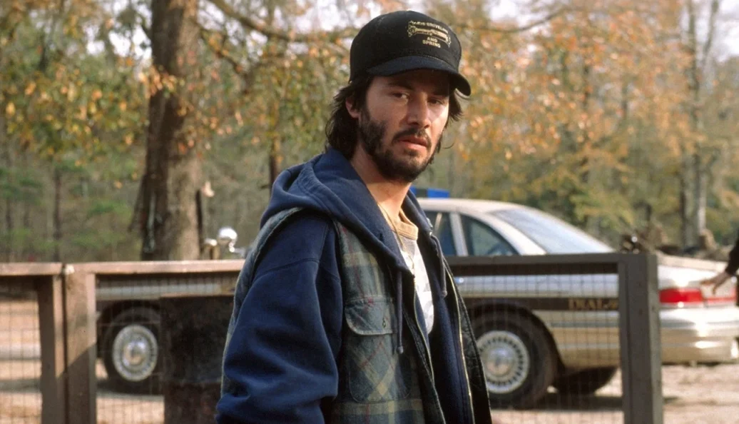 Keanu Reeves: Fotografia de Keanu Reeves em uma cena do filme The Watcher, usando um boné preto e casaco azul.