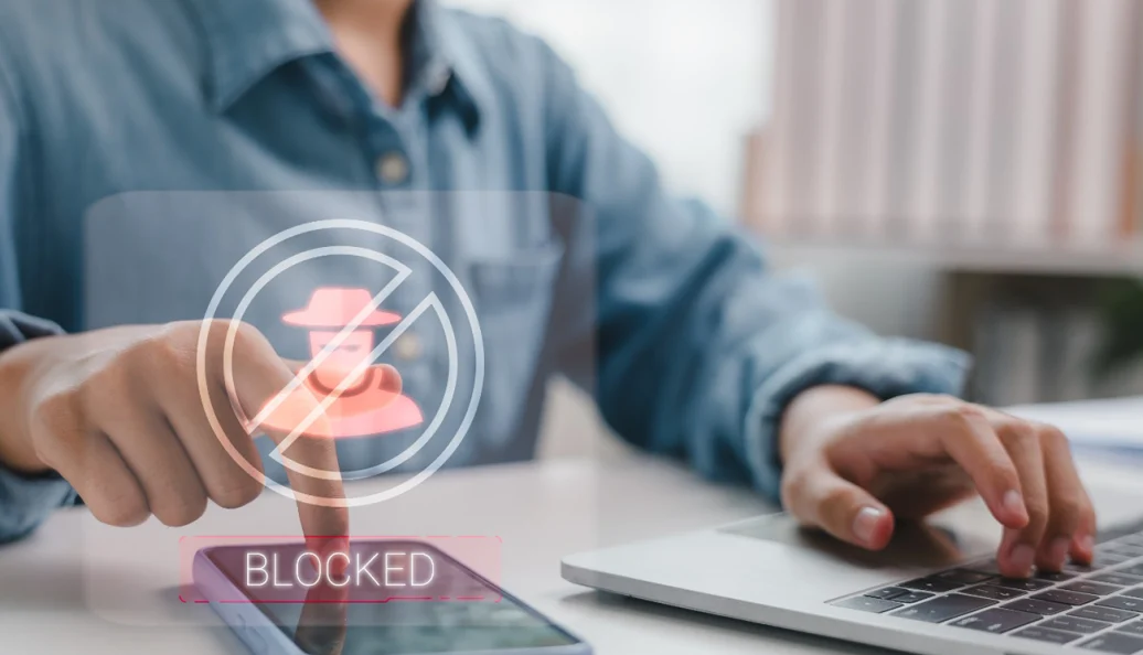 unknown: Uma pessoa utilizando um smartphone e um notebook com um gráfico sobreposto de segurança digital exibindo um ícone de hacker bloqueado.
