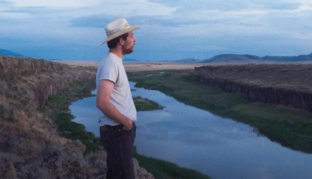 unknown: Um homem de chapéu branco e camiseta cinza observando uma paisagem de cânion com um rio ao fundo durante o crepúsculo.