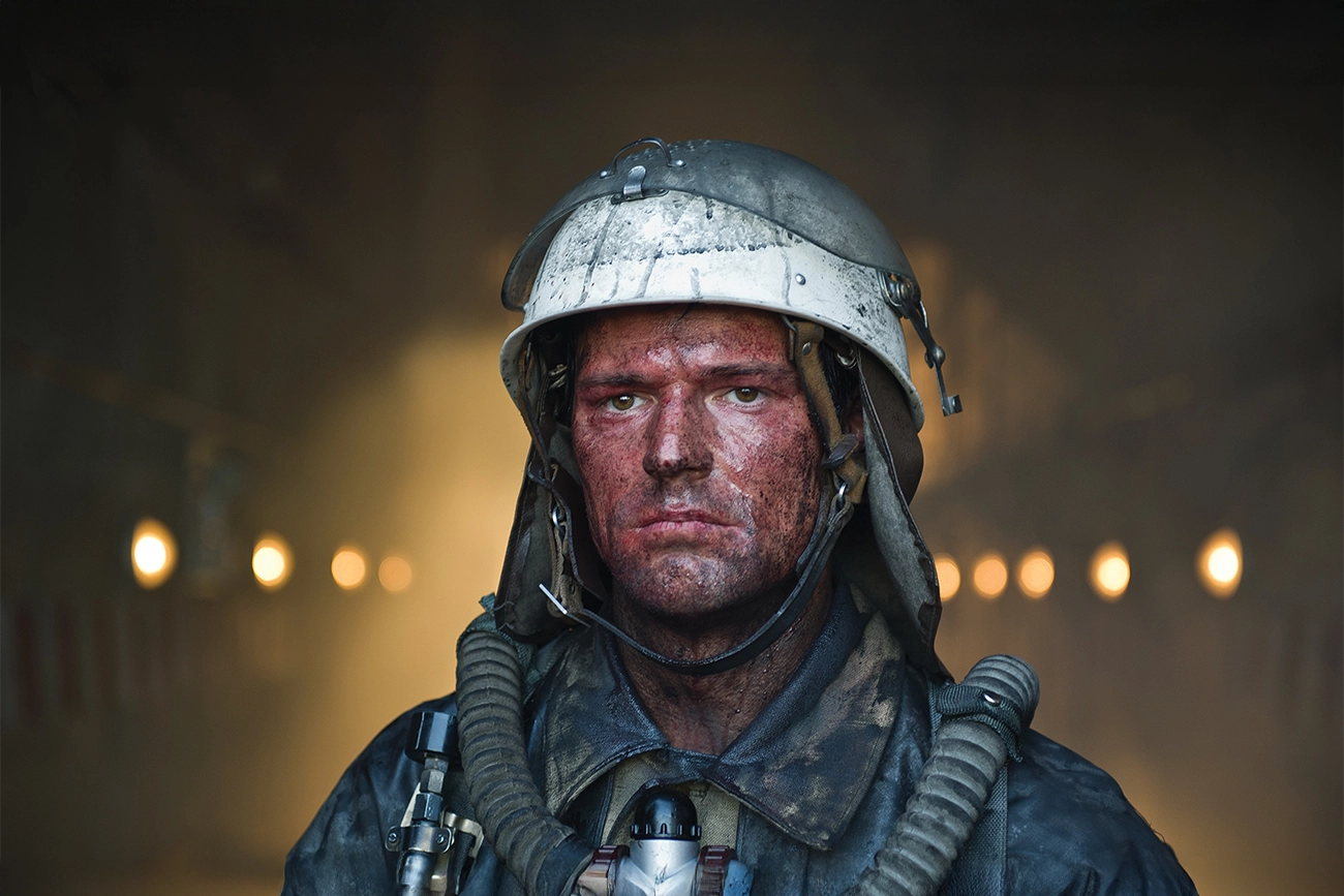 unknown: Retrato de um homem com o rosto sujo de fuligem, usando capacete e equipamento de proteção em um túnel ou ambiente industrial.