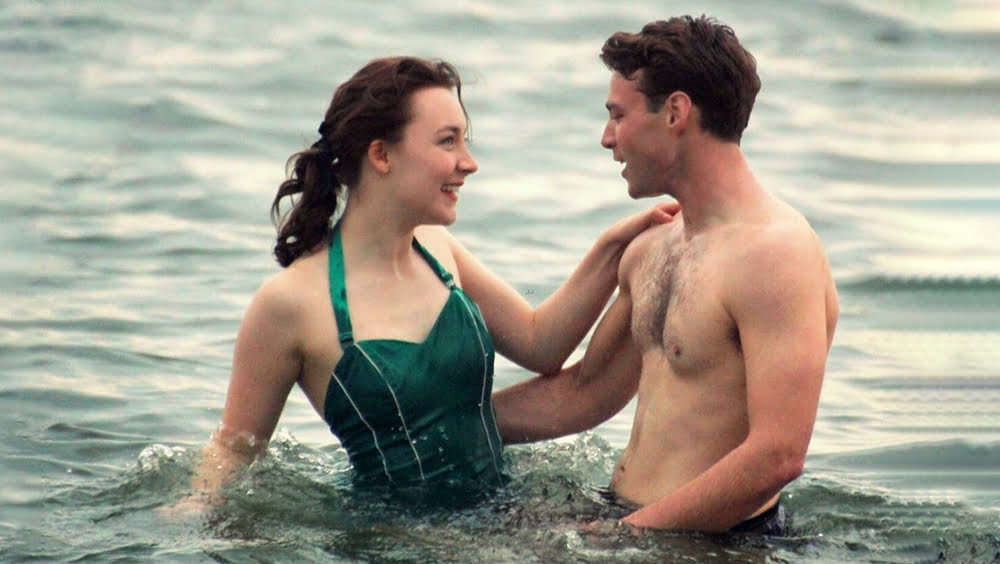 Saoirse Ronan e Emory Cohen: Uma cena do filme Brooklyn (2015) mostrando um casal no mar, com a mulher em um maiô verde e o homem sem camisa.