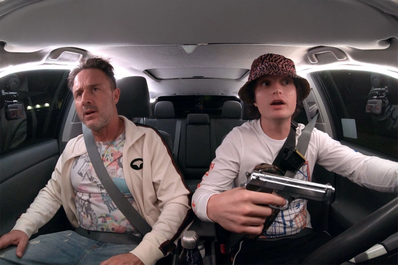 David Arquette: Uma cena de filme dentro de um carro com o motorista segurando uma arma e um passageiro ao lado, ambos com expressões de surpresa ou choque.
