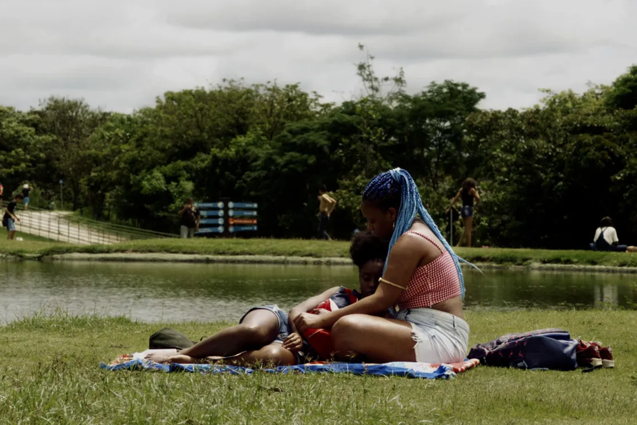 unknown: Duas pessoas sentadas em uma toalha na grama em frente a um lago em um parque.