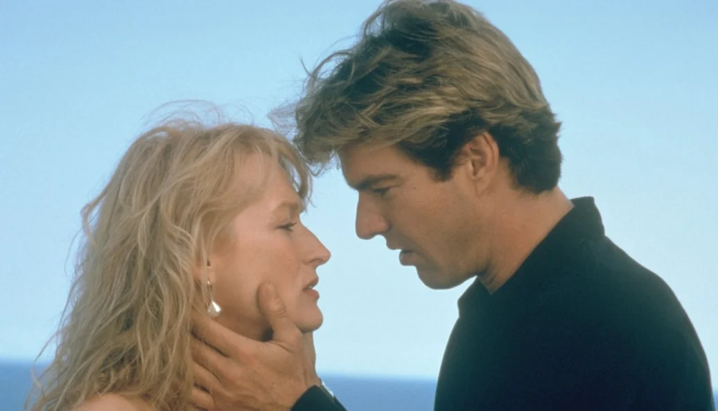 Meryl Streep, Pierce Brosnan: Uma cena romântica de filme onde um homem segura o rosto de uma mulher loira, prestes a se beijarem.
