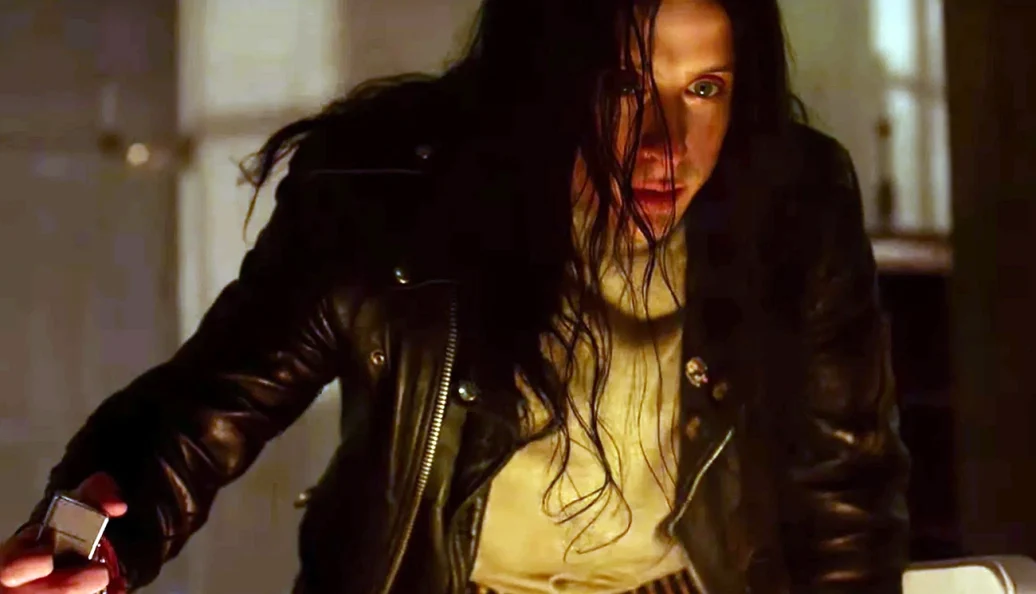 Noomi Rapace (como Lisbeth Salander): Uma pessoa com cabelos longos e escuros, vestindo uma jaqueta de couro preta e segurando um objeto metálico, em uma cena de filme.