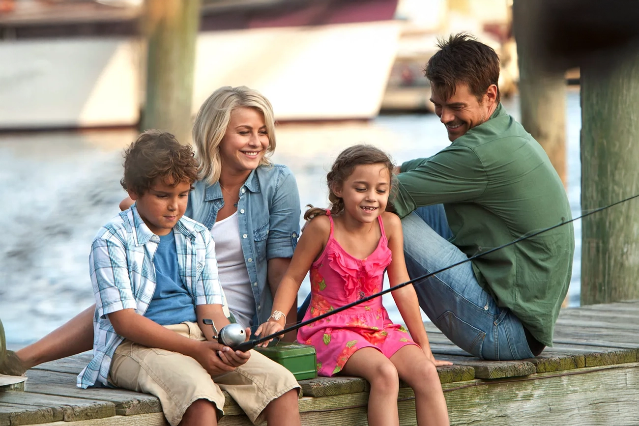 Julianne Hough, Josh Duhamel: Uma família composta por dois adultos e duas crianças sentados em um píer de madeira, com o menino segurando uma vara de pescar. Cena do filme 'Safe Haven' (Um Porto Seguro).
