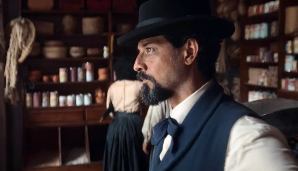 Antônio Haddad: Homem com barba e chapéu preto em um ambiente que remete ao século XIX, possivelmente uma venda ou armazém, em cena de filme ou série.