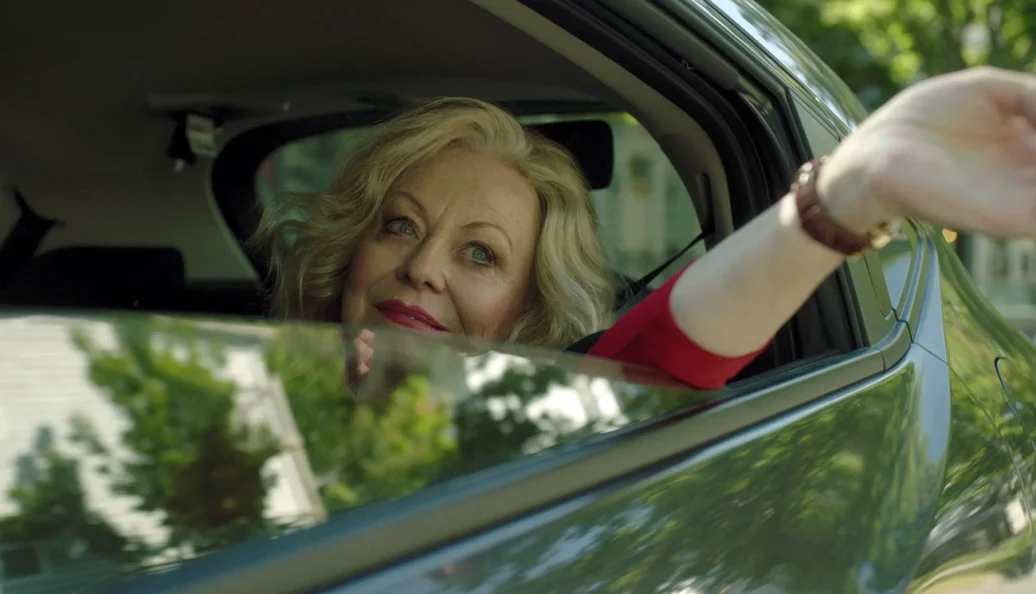 Jacki Weaver: Uma mulher loira sorrindo e acenando pela janela traseira de um carro.