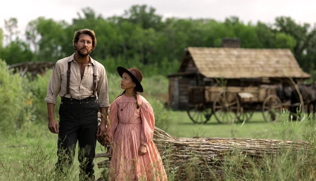 Jacob Lofland, Cailee Spaeny: Um homem e uma jovem em trajes de época do século XIX em um ambiente rural com uma cabana de madeira e carroça ao fundo.
