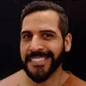João Moreira: Retrato de um homem sorridente com barba, cabelo curto e fundo preto.