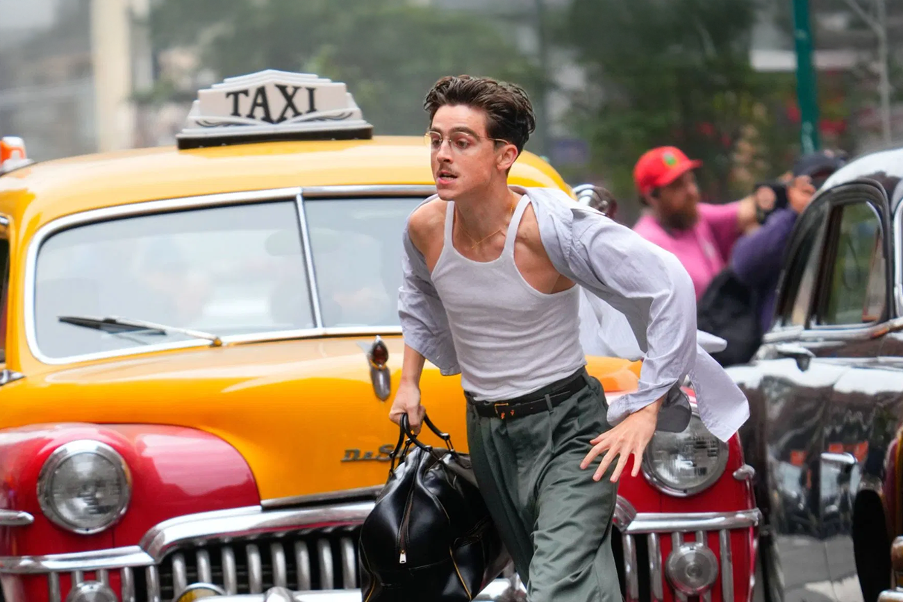 Josh O'Connor: O ator Josh O'Connor correndo em uma rua movimentada, vestindo regata e segurando uma mala, em frente a um táxi antigo amarelo e vermelho.
