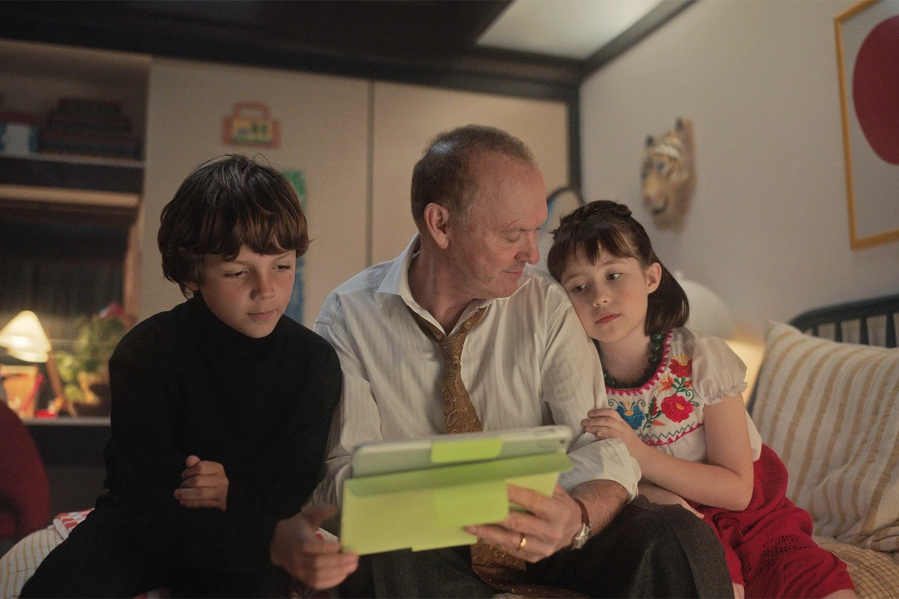 Michael Keaton: Uma cena do filme Birdman mostrada Michael Keaton sentado em uma cama com duas crianças enquanto olham para um tablet.