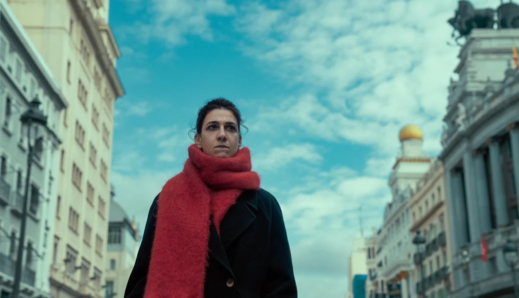 Antonia Scott (Victoria Luengo): Uma mulher usando um casaco preto e um cachecol vermelho felpudo, em pé em uma rua da cidade com prédios clássicos e céu nublado ao fundo.