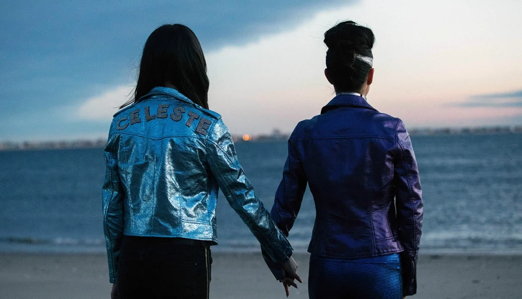 Natalie Portman, Raffey Cassidy: Duas mulheres de costas segurando as mãos em uma praia, uma delas usando uma jaqueta prateada com o nome 'CELESTE'. Cena do filme Vox Lux.