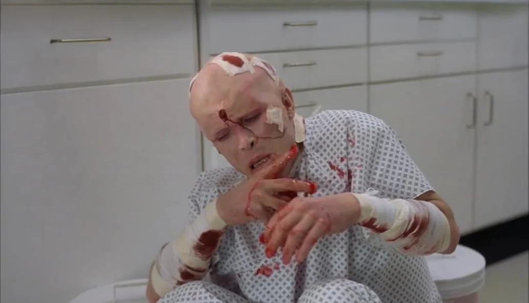 David Bowie: Uma cena de filme mostrando um homem ferido e ensanguentado com bandagens, sentado no chão de um ambiente hospitalar.