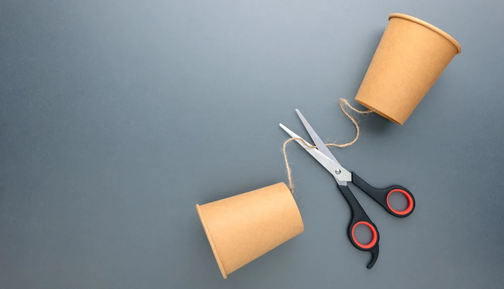 Dois copos de papel conectados por um barbante que está sendo cortado por uma tesoura, simbolizando quebra de comunicação.