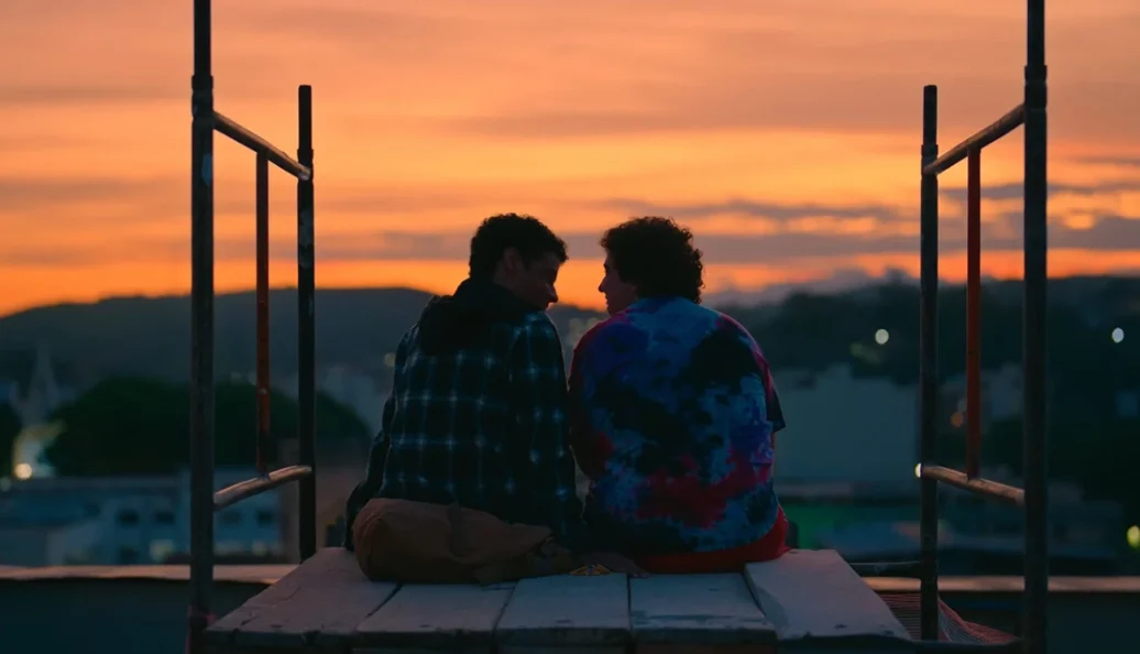 Gabriel Leone, Maurício Destri: Dois jovens sentados em um andaime de madeira no topo de um prédio, observando o pôr do sol em uma área urbana.