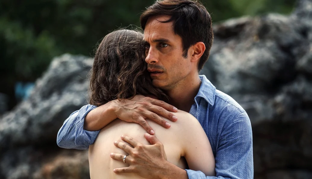 Gael García Bernal: Um homem com expressão de preocupação ou choque abraçando uma pessoa de costas em um ambiente externo com rochas ao fundo.