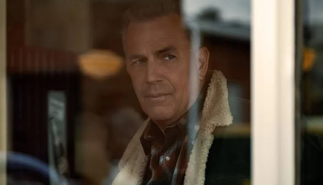 Kevin Costner: Um homem de meia-idade com expressão pensativa, usando uma jaqueta com gola de lã, visto através do reflexo de uma janela. Cena da série Yellowstone.