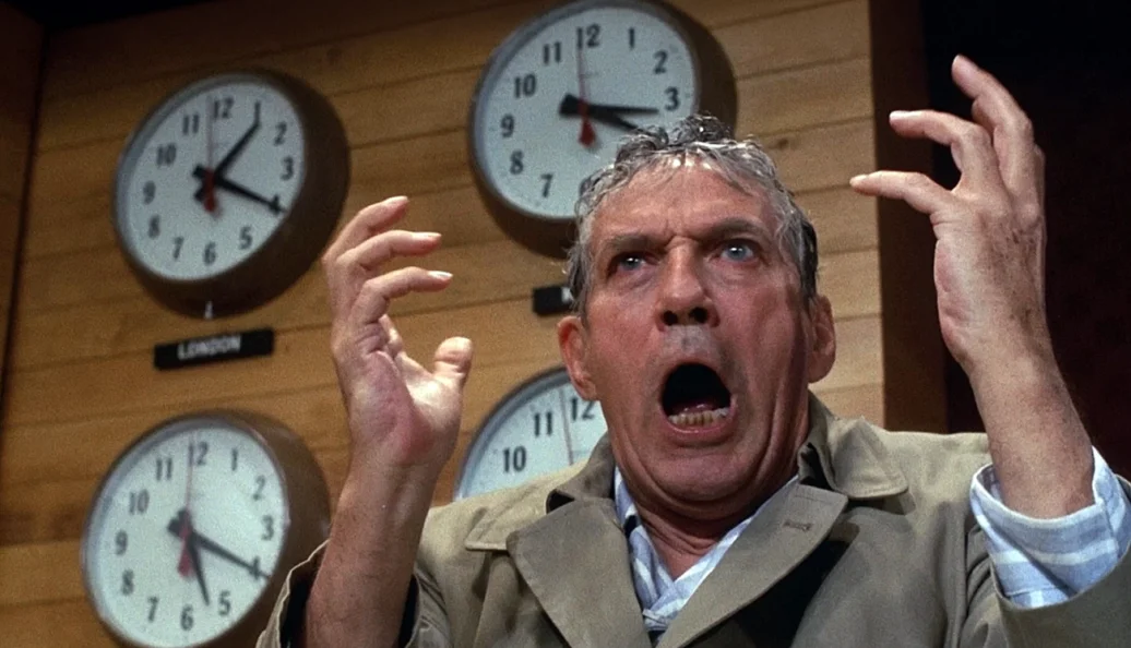 Peter Finch: Homem gritando com as mãos levantadas em frente a vários relógios de parede, cena do filme Network (1976)